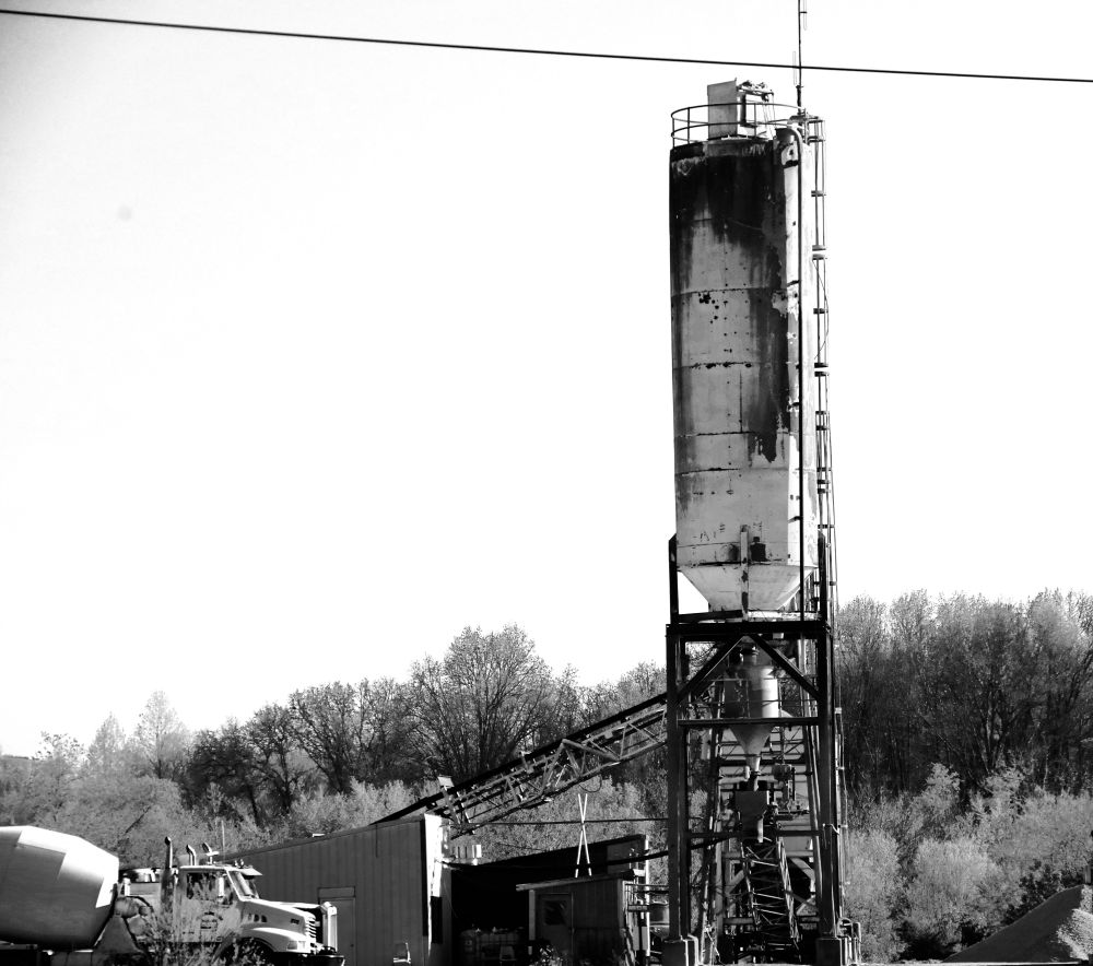 A black and white photograph of a cement silo in front of a wooded area. A sloped conveyor belt leads towards the ground, and a cement mixing truck waits near the foot of the belt. 
