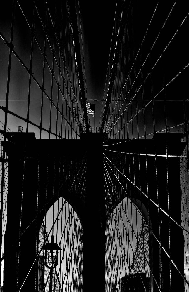A black and white solarized image of a huge arched suspension bridge tower, taken along the axis of the bridge. A grid of steel cablework radiates from the tower, reflecting sunlight, and an American flag waves atop it. 