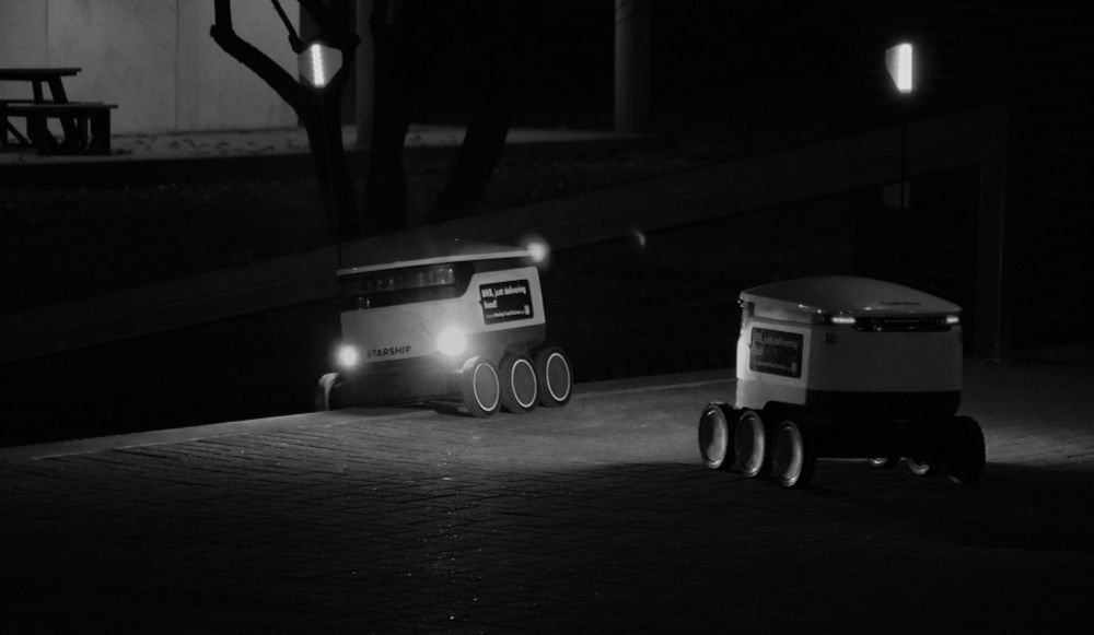 A black and white photograph of two six-wheeled delivery robots at night. One robot's headlights can be seen as it sits tilted on the edge of the top step of a flight of stairs. The second robot illuminates the stuck robot with its headlights as it faces away from the camera. 