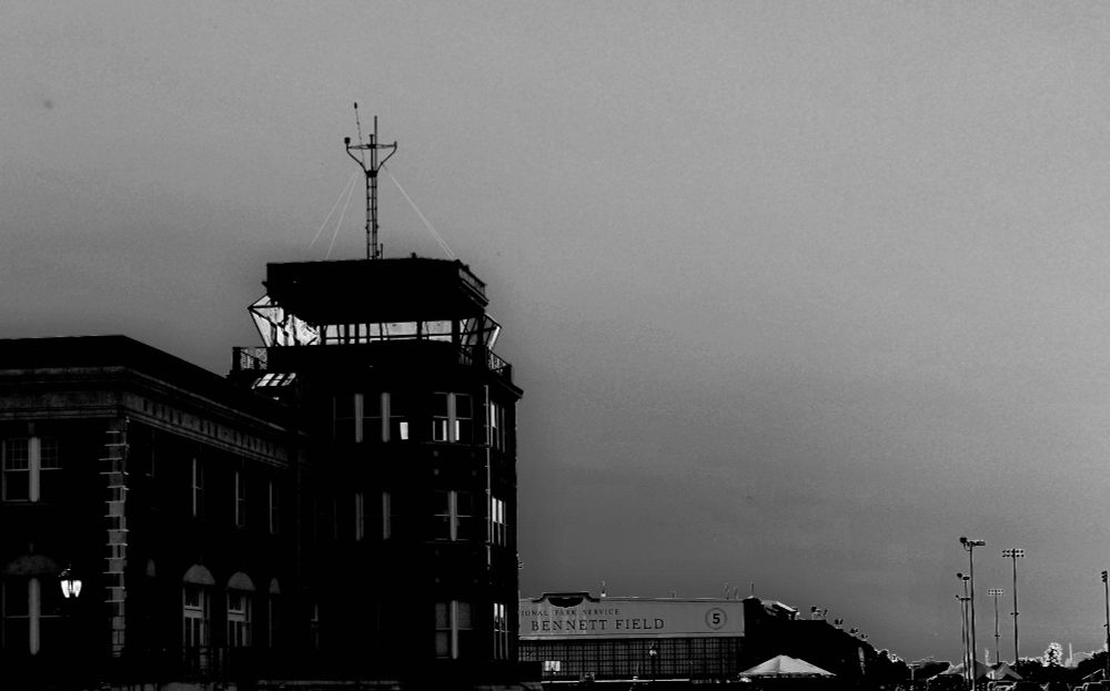 A black and white solarized photograph of a two-story brick building with an attached four-story air traffic control tower topped by a radio mast. In the background an aircraft hanger can be seen with the words "Bennet Field" visible above the doors.