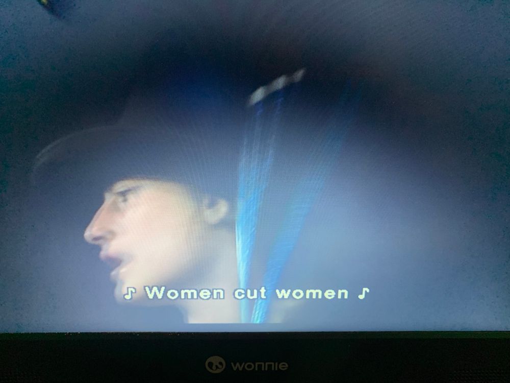 Blurry image of a woman holding a drum stick. Subtitle reads, “Women cut women” with music signs to show you it’s a song