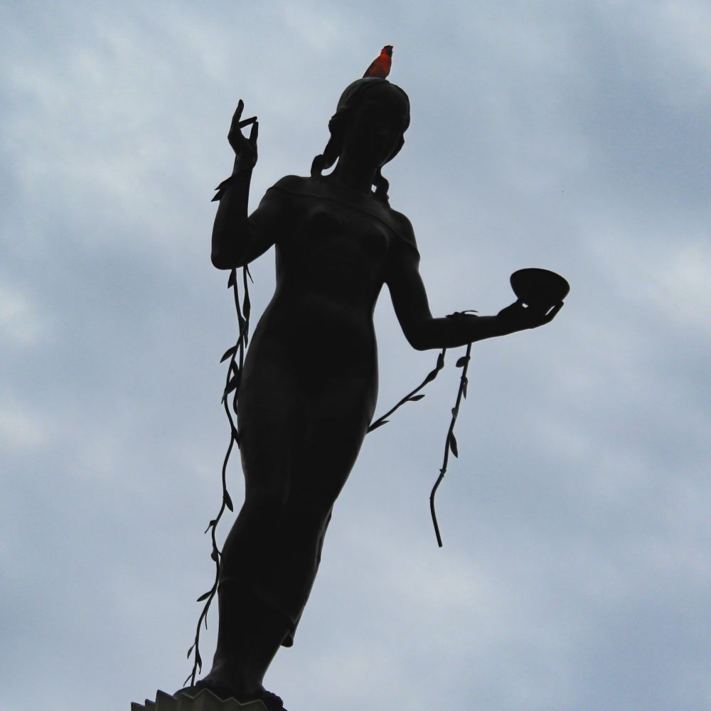 Silhouetted against a cloudy, grey sky is the statue of a woman facing the lens. Details on the statue are hidden by shadow. She has both arms bent, her right hand raised with her thumb and pointer finger touching, and her left hand holding a bowl. A vine with small leaves is draped over her arms. On the statue’s head is a male cardinal, some of his bright red color popping even though he is also being silhouetted by the muted light.
This photo was taken by Taralei Griffin at Allerton Park in Monticello, Illinois in June 2021.
