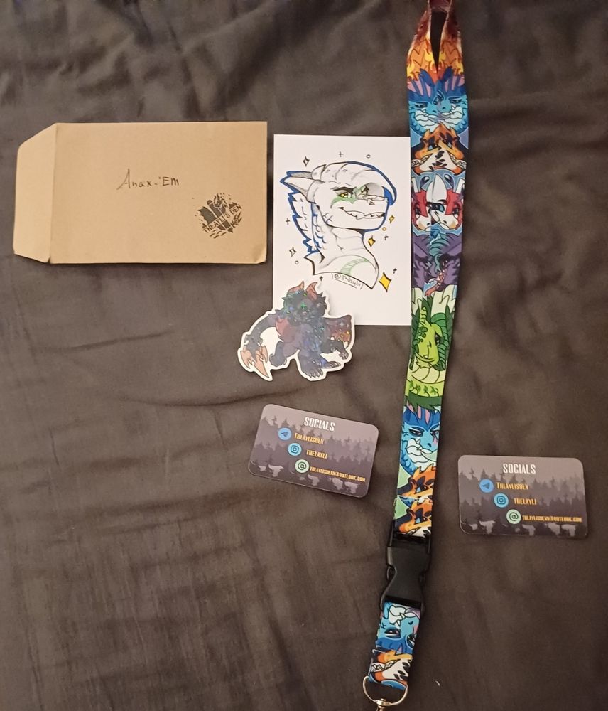 An envelope adressed to "Anax'em" with a sketch portrait of Anax'em - a dragon smiling. A holographic sticker of Thlayli - a dark purple dragon, a lanyard featuring a variety of dragons (with the keys cut off the picture for safety reasons) and socials of Thayli!