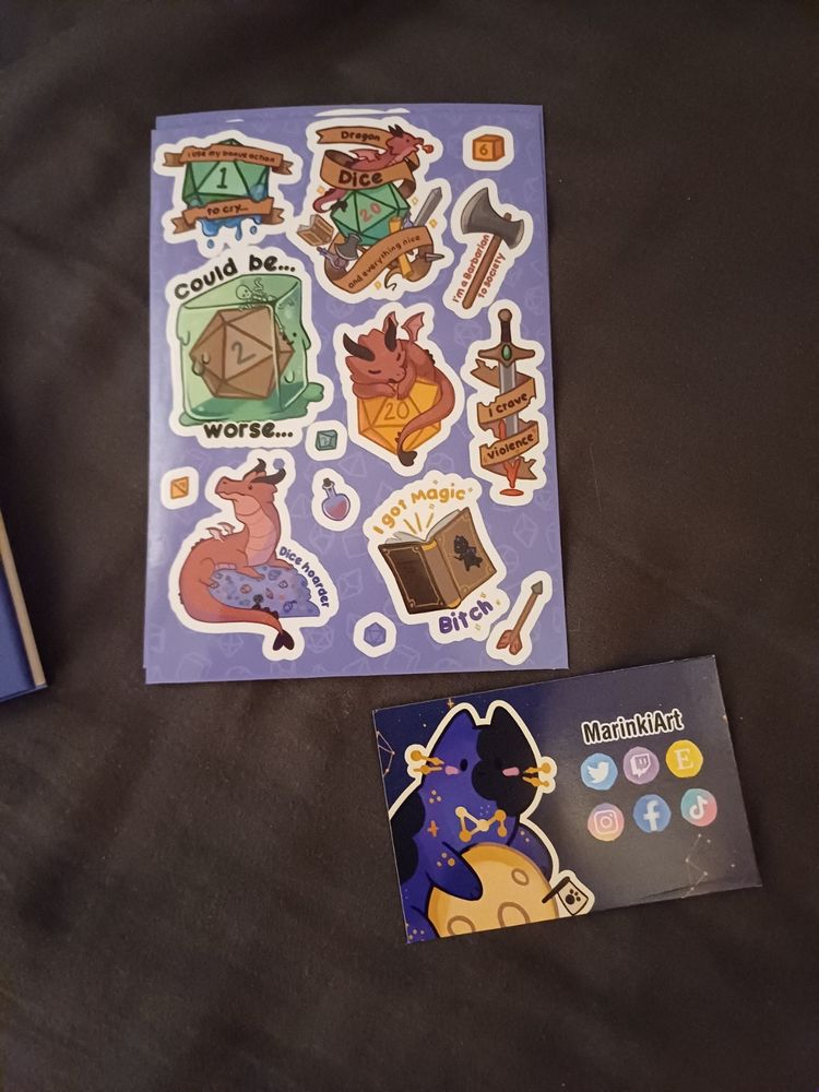 A bunch of stickers that says in order "I use my bonus action to cry" overlayed over a Nat 1, "Dragon Dice and everything nice" with a hoard of stuff, a D6, an Axe with "I'm a barbarian to society", "Could be worse" with a Nat 2 stuck inside of a slime, a dragon sleeping on a D20, "I crave violence" surrounding on a bleeding sword, a d4?, Dragon hoarding dice with the text "Dice hoarder", a potion, a small d20, A spell book with the text "I got Magic Bitch" and an arrow. Also a card with MarikiArt's socials.