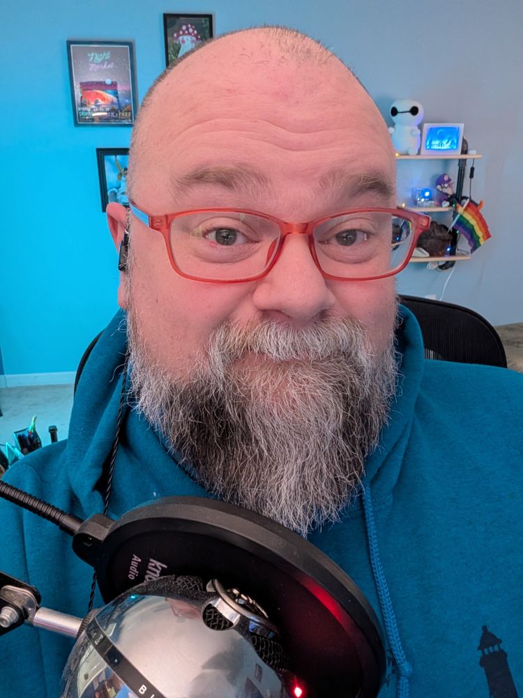 Smiling bald bearded person wearing red glasses in front of a microphone