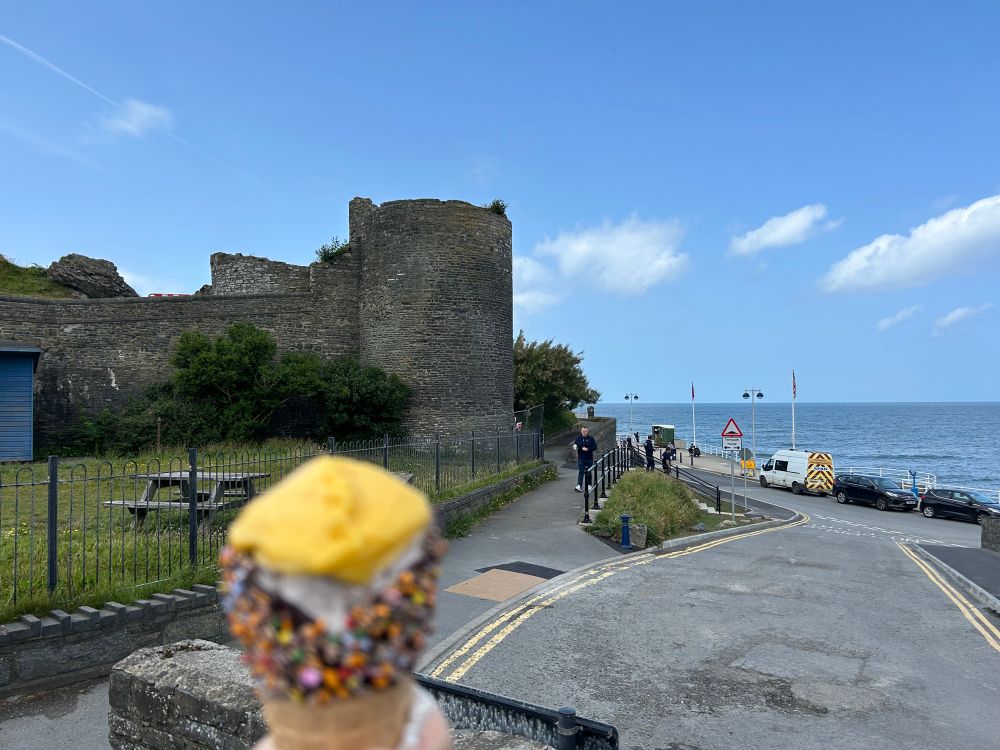 A photo from a first person perspective of a ruined castle next to the sea, between which runs a road. In the foreground, out of focus, is a large ice cream cone with grey and white and yellow ice cream.