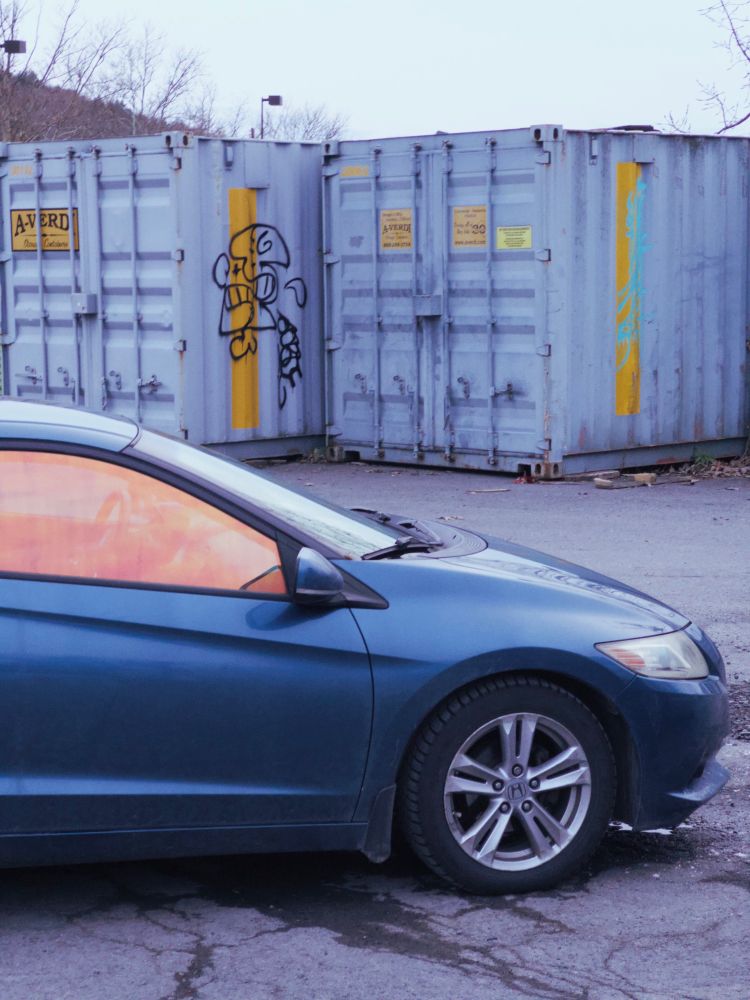 A couple of blue and yellow shipping containers foregrounded by a blue honda with orange tinted windows