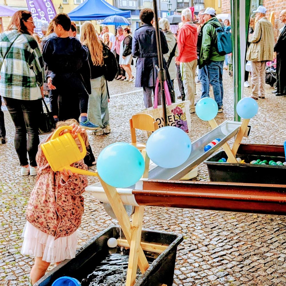 Kind gießt Wasser mit Gießkanne an einem Wasserspielplatz. Im Hintergrund stehen Menschen u.a. ein Pfarrer.