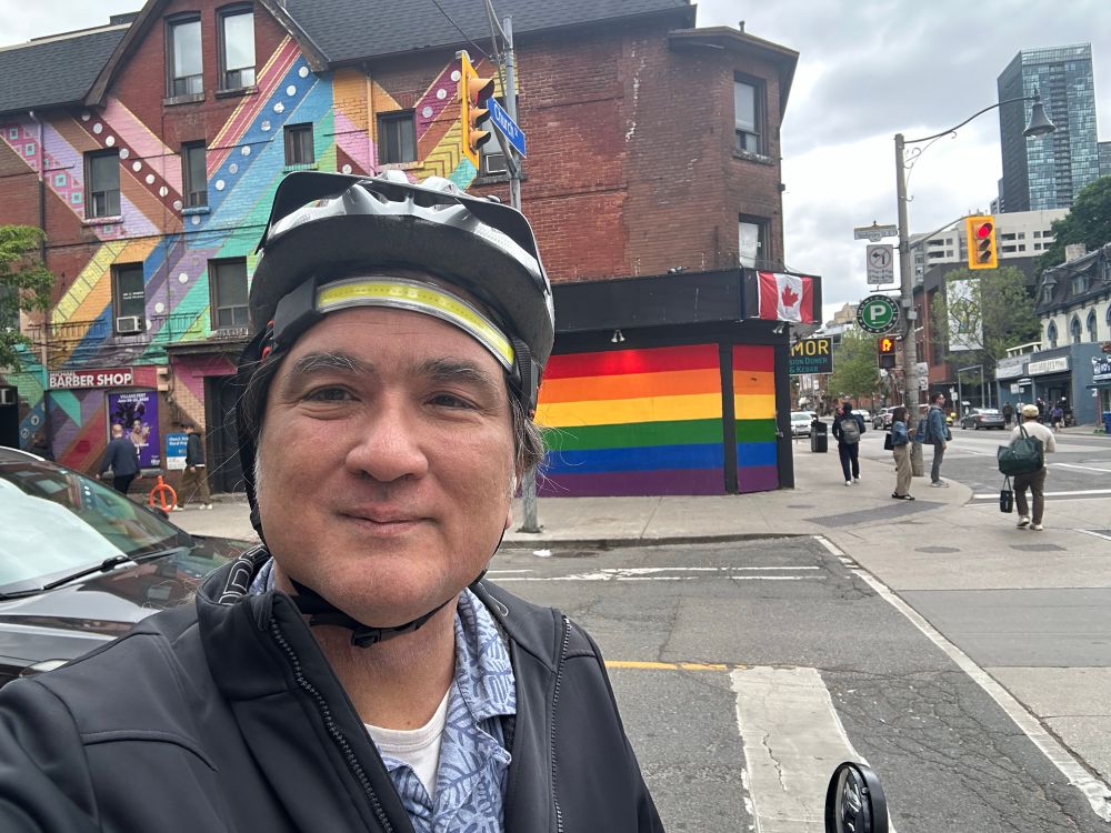 John poses on his bike wearing a helmet and LED headlamp, in front of a rainbow Pride mural at Church and Wellesley, the heart of the Village in downtown Toronto