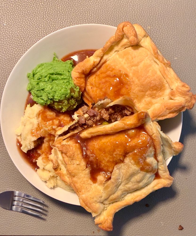 Das Bild zeigt einen weißen Teller auf einer strukturierten, hellgrauen Oberfläche.
Auf dem Teller liegen zwei große, rechteckige Blätterteigpasteten mit leicht aufgeplatzten, goldbraunen Deckeln. Eine der Pasteten ist an einer Seite geöffnet, sodass eine Füllung aus braun gebratenem Sojahack und etwas Zwiebel sichtbar ist. Zwischen und unter den Pasteten befindet sich bräunliche Soße.

Links auf dem Teller liegen zwei Beilagen: unten ein heller, cremiger Haufen Kartoffelpüree, leicht mit Soße bedeckt; darüber ein kompakter, kräftig grüner Klecks Erbsenpüree.

Am unteren linken Bildrand liegt eine silberne Gabel, teilweise sichtbar.