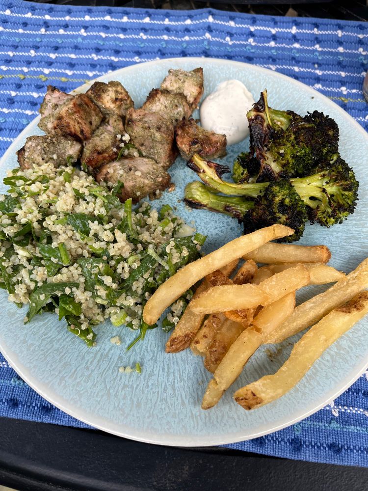 A plate with nicely browned grilled jerk pork kebab chunks, grilled charred broccoli, quinoa spinach salad, and French fries 