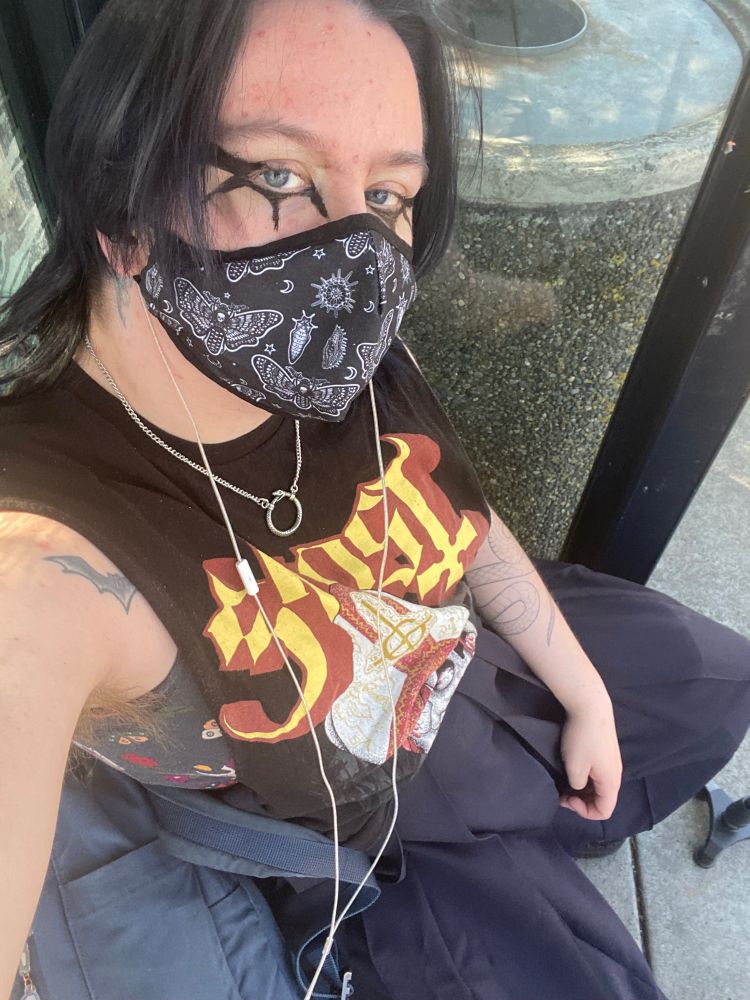 Dorian at a bus stop. dressed in a muscle band tee, hakama, moth mask, oroboros necklace and looking at the camera.