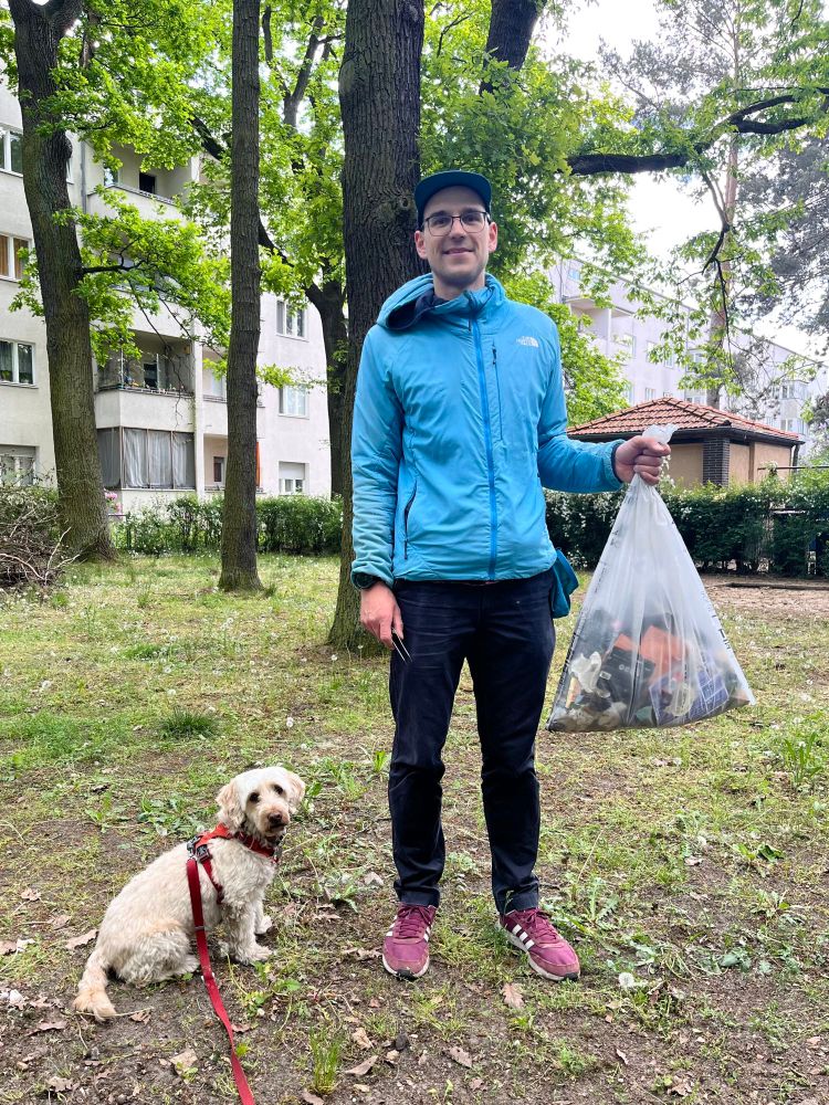Ein junger Mann steht vor einem Haus. In der linken Hand hält er einen transparenten Müllsack, der zu einem Großteil mit Müll gefüllt ist. Neben ihm sitzt ein Hund. 
