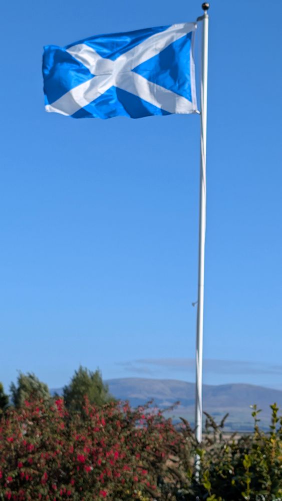 Saltire flying from a flagpole with hills in the distance.
