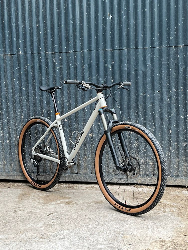 Picture of HROK hardtail 29er mountain bike shot at an angle in front of a farmyard corrugated metal door. Bike is painted a light stone colour and fitted with tan wall tyres. 