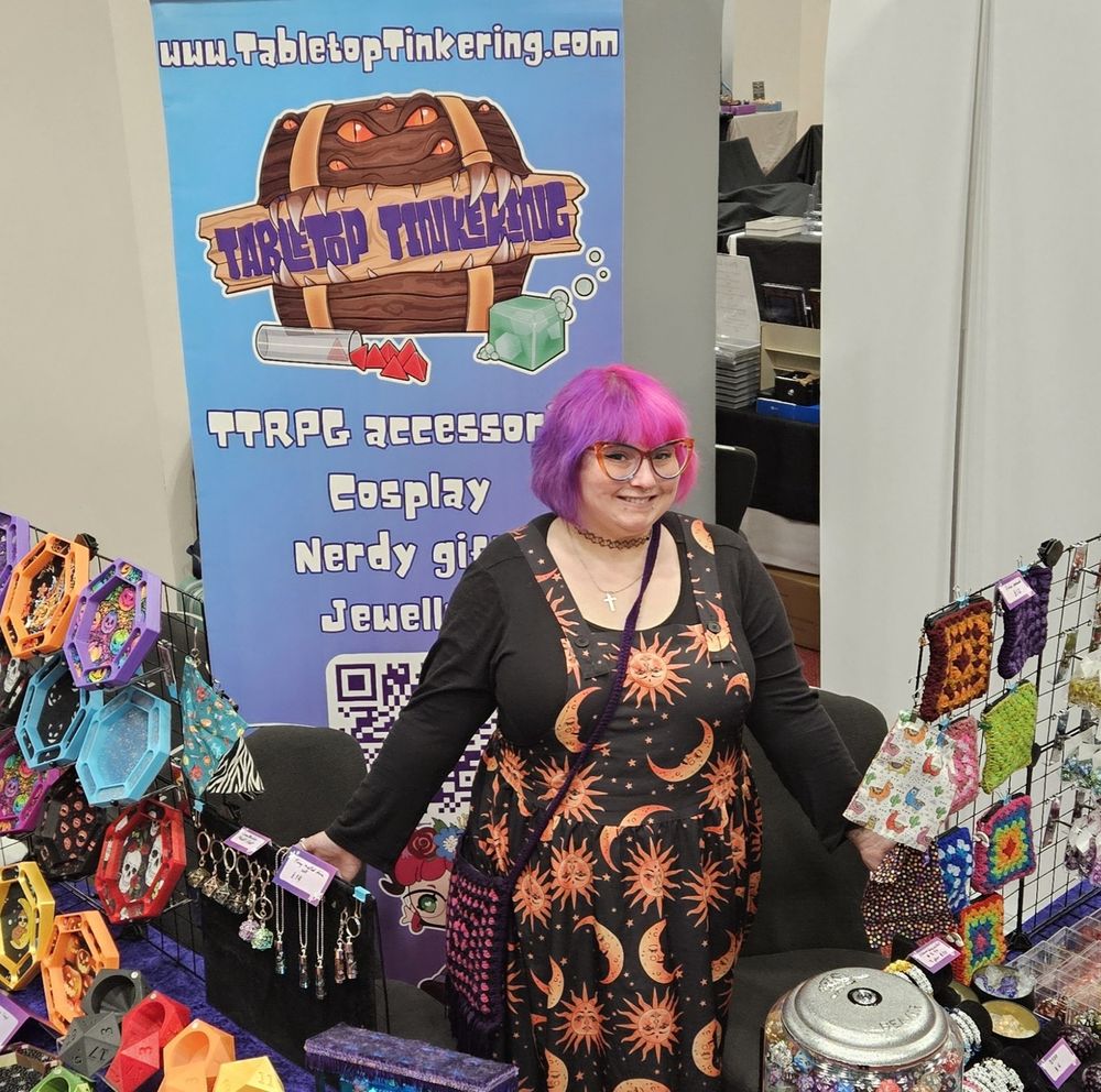 A person with pink and purple hair and a dress covered in moons and sins standing behind a stall