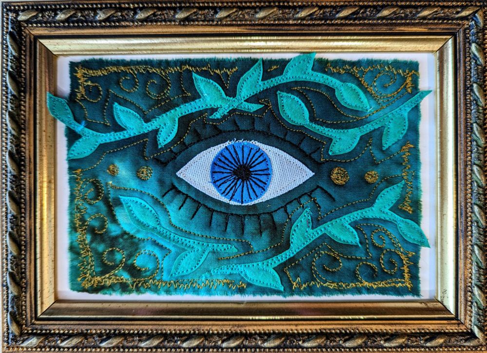 a small, quilted piece of art featuring an evil eye on hand-dyed blue fabric with an intricate frame 