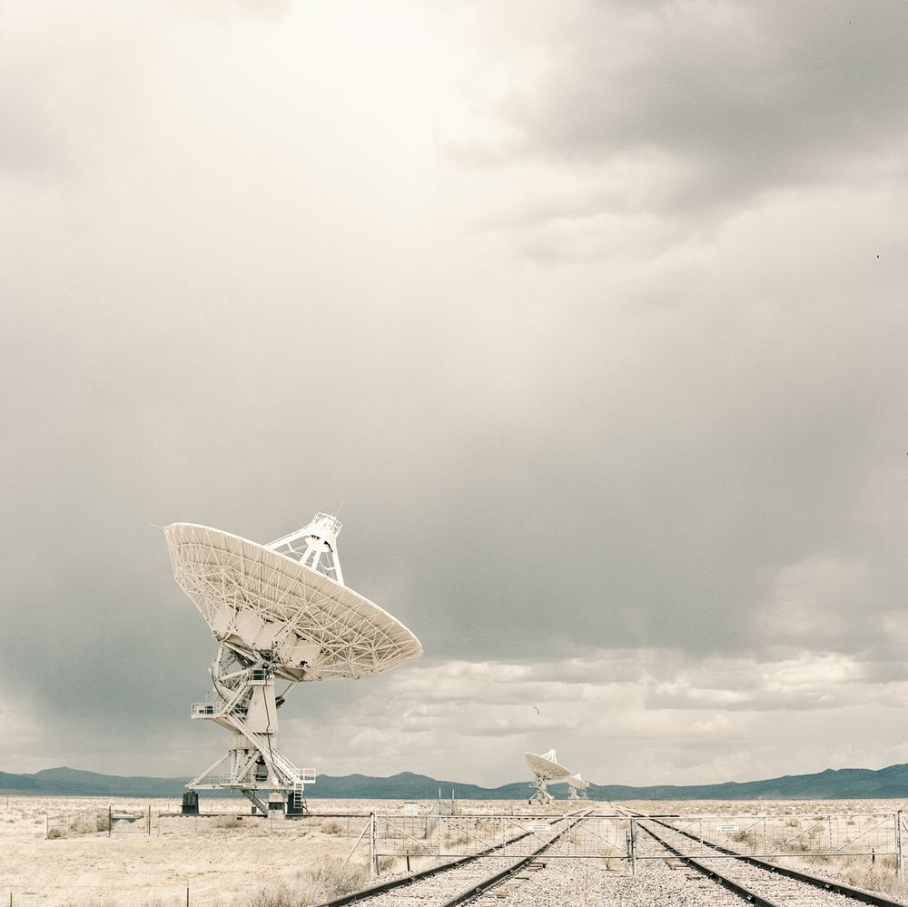 This image shows few telescopes of the Very Large Array in New Mexico. The sky is cloudy with rays of sun shining through and illuminating the foreground telescope. Two railway tracks enter the image at the lower right.