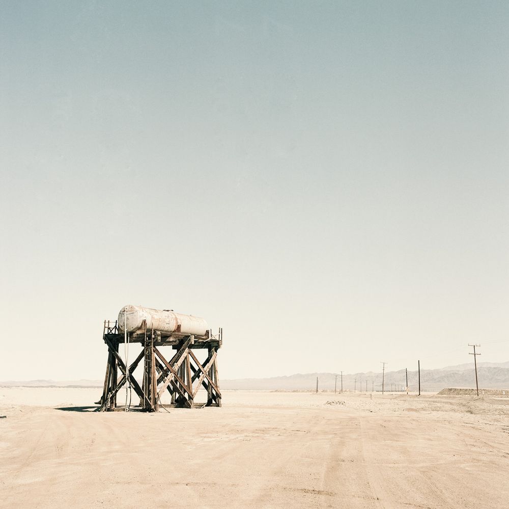 Image shows a flat, desert like area. At the right one can sense a street with electricity poles. The main subject is in the left of the image. It is a tank, maybe water tank on a wooden construction. The tank is in about 5-7 meter hight. The mood of the image is kind of dystopian as it seems an abandoned place.