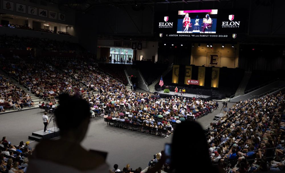 Attendees at an Elon University event seated in a large indoor arena, watching a speaker on stage, with the stage displayed on two large screens.