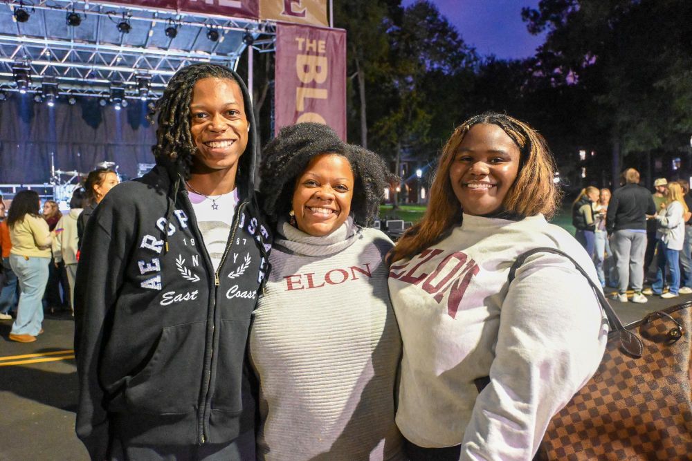 Three individuals smiling at an Elon University event, standing in front of a stage with "The BL" signage, during twilight. Two are wearing university apparel.
