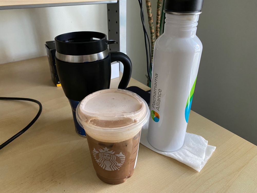 A photo of three beverages on a light wood desk: a Starbucks cold brew, a Pachamama Alliance metal water bottle, and a blue and black travel mug