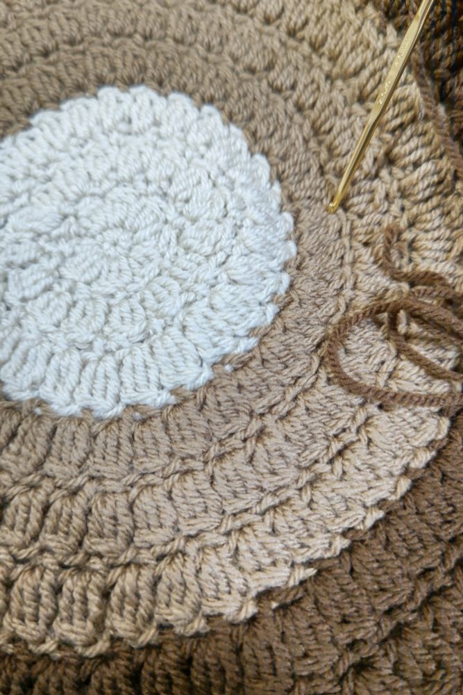 かぎ針編みで編まれた円形の座布団が写っている。座布団の配色は内側から順に白色、うす茶色、茶色のグラデーションで、金色のかぎ針と茶色の毛糸の端が写り込んでいる。
