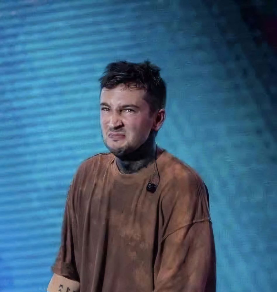 Tyler Joseph from twenty one pilots has a brown shirt, black neck paint, his in-ear out, making a stank face in front of a blue background 