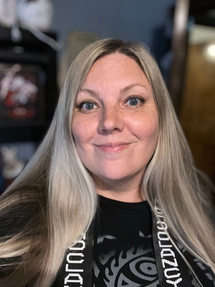 A blonde woman smiles at the camera with a black tribal shirt and black and white stole that says Clancy