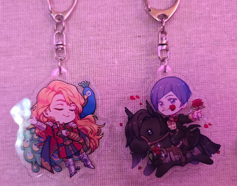 A keychain featuring a smug war Ferdinand in front of a peacock and a keychain featuring romantic academy Lorenz riding a horse with roses in his hand and mouth