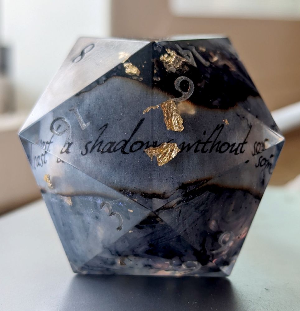 a giant d20 with black and silvery swirls, flakes of gold foil, and a slip of scorched paper that reads "You can't cast a shadow without some light."