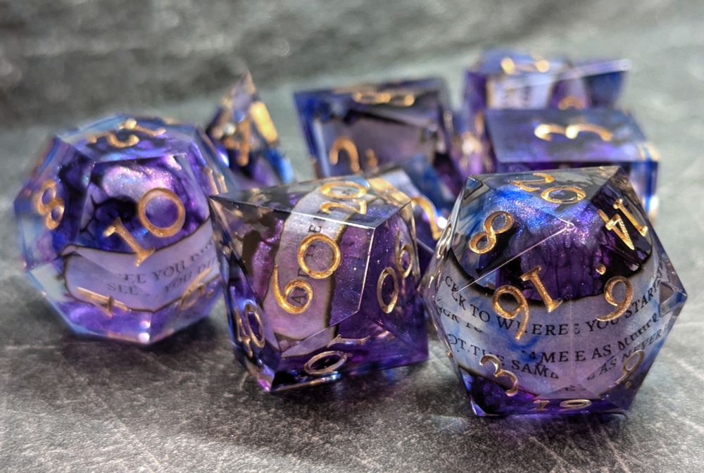 A set of polyhedral dice for tabletop games. The d20 is in foreground. They have soft shades of purple and blue, cut through with veins of darker purple, and contain small burned slips of paper with printed words on them. They are inked in gold.