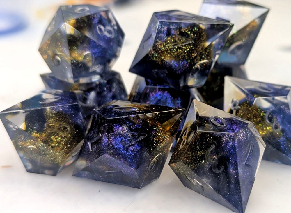 A set of elongated 10-sided dice, made of clear resin with swirls and clouds of colorshifting mica in shades of blue-violet, gold-green, and holographic black.
