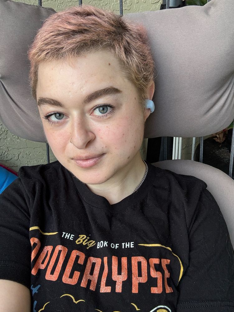 a selfie of Emmy, a feminine person with short pink hair, wearing a pair of baby blue open ear headphones. This type of headphone clips to the outside of your ear rather than sitting in the ear canal. 