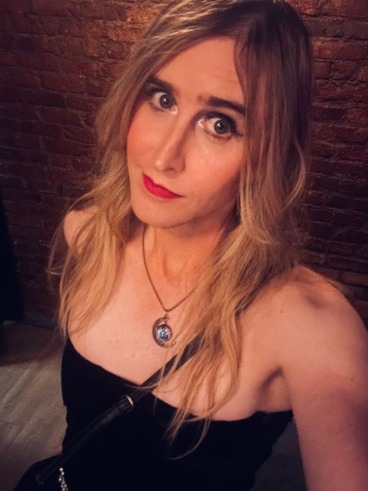 emma in a short black dress, cosmic necklace with a blue and purple galaxy. her blonde hair is draped down her shoulders and she's looking up at the camera with a smirk and her big green eyes staring into your gay little soul real big