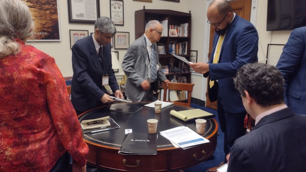 Adults in business attire are preparing to sit at a round table in an office just before the start of a meeting.