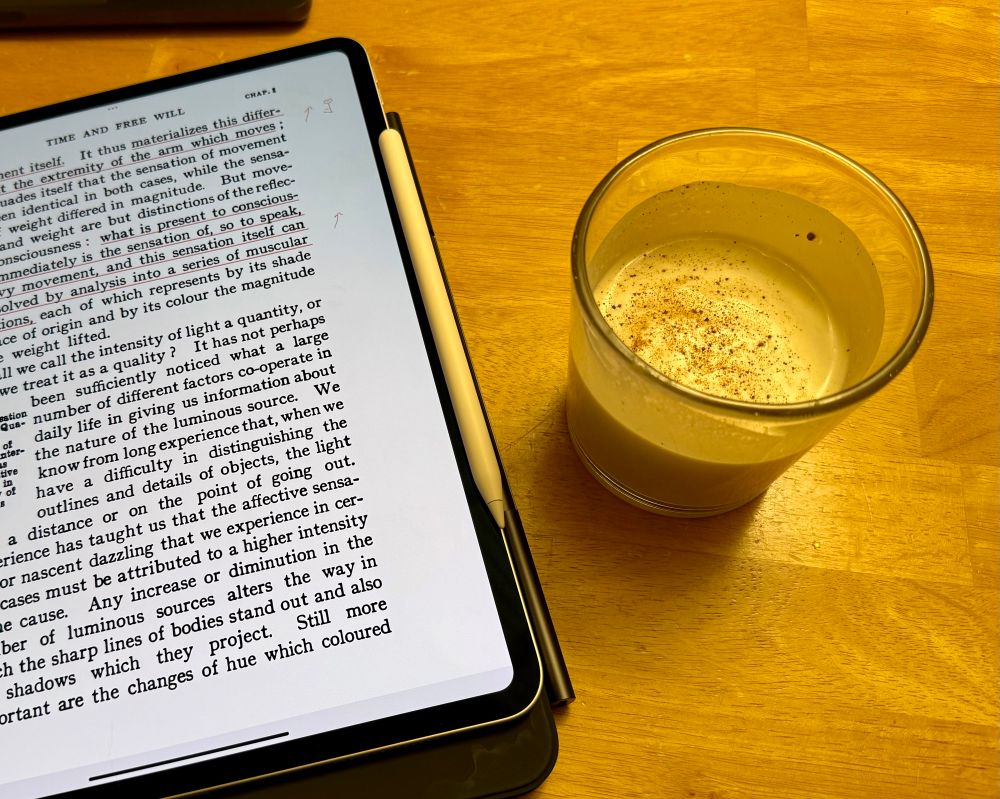 An iPad showing a page of Bergson’s Time and Free Will and a rocks tumbler filled with eggnog and nutmeg, both sitting on a blond wood table. 