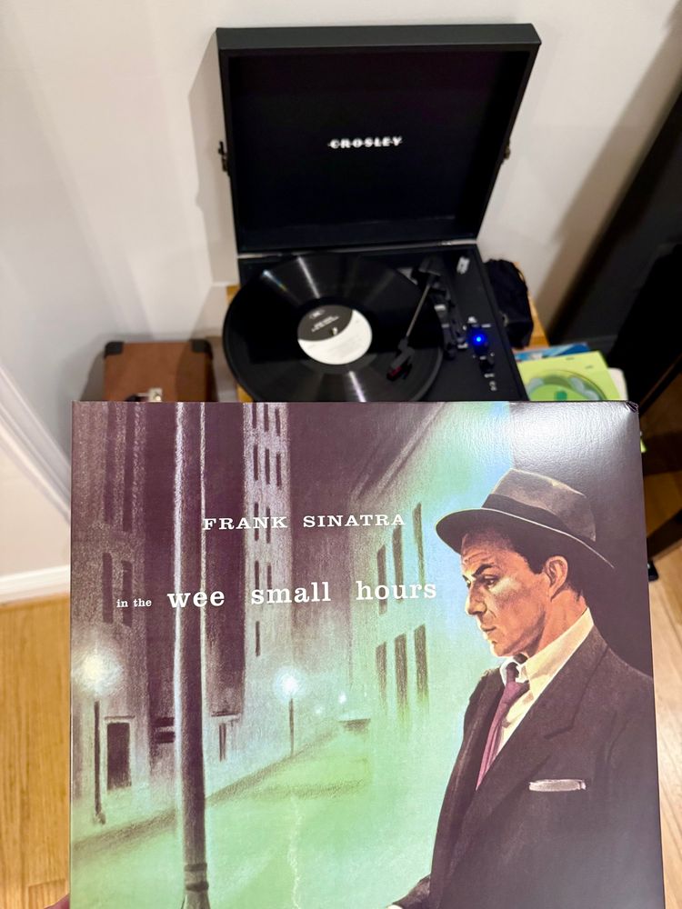 Cover of Frank Sinatra’s album, “In the Wee Small Hours,” depicting a painting of a young Frank Sinatra wearing a grey suit, loosened tie, and fedora, with a night city street behind him. Behind the album cover is a record spinning on a record player. 
