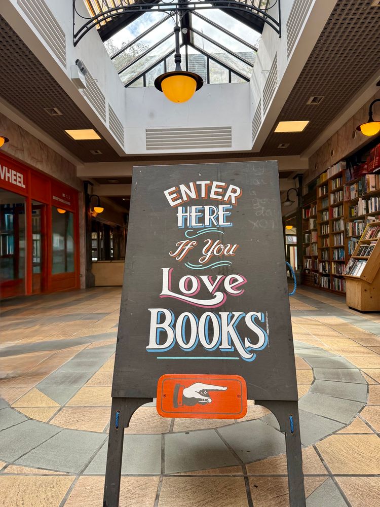 A sign reading “Enter Here If You Love Books” with a hand pointing right, sitting on a tile floor with books on shelves behind it to the right. 