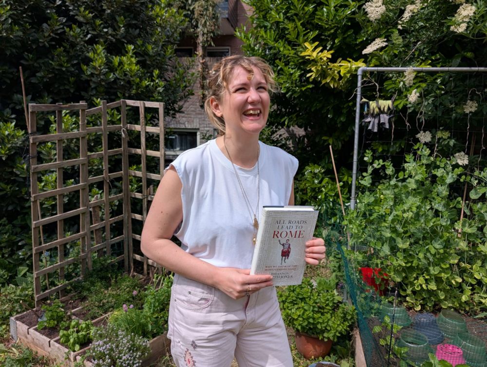 Photo of the author, a white woman with blonde hair, in a plain white t-shirt and pink, embroidered jeans, standing in a sunny garden, laughing, and holding the book All Roads Lead To Rome