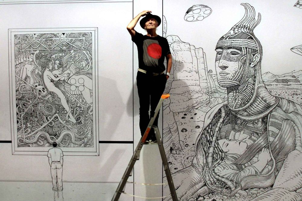 Artist Jean Moebius Giraud standing on a ladder in front of a mural of his black and white artwork. It looks to be the preparations for an art exhibit.