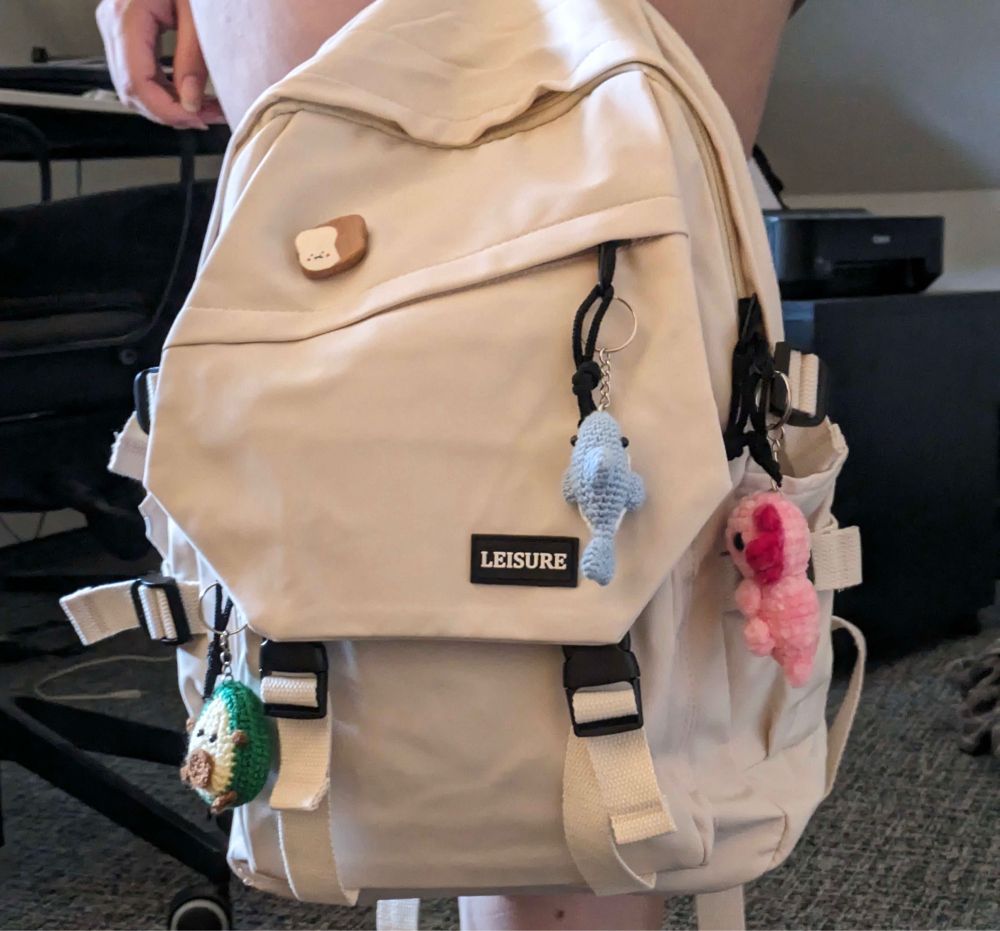 A white backpack with a shark, an avocado, an axolotl keychain and a pin