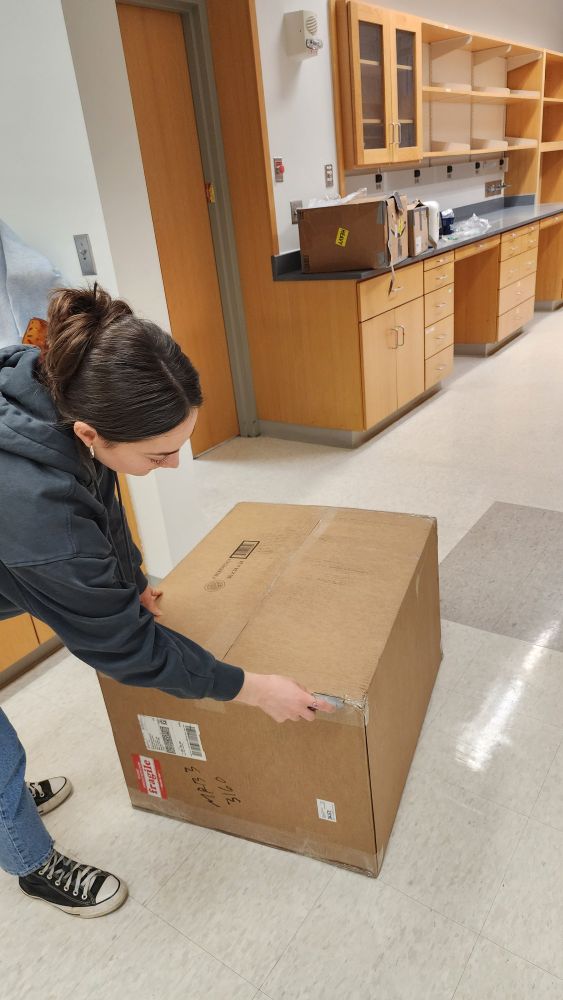 Woman in dark gray hoodie using box cutters to open up a medium sized brown box on a white tiled floor