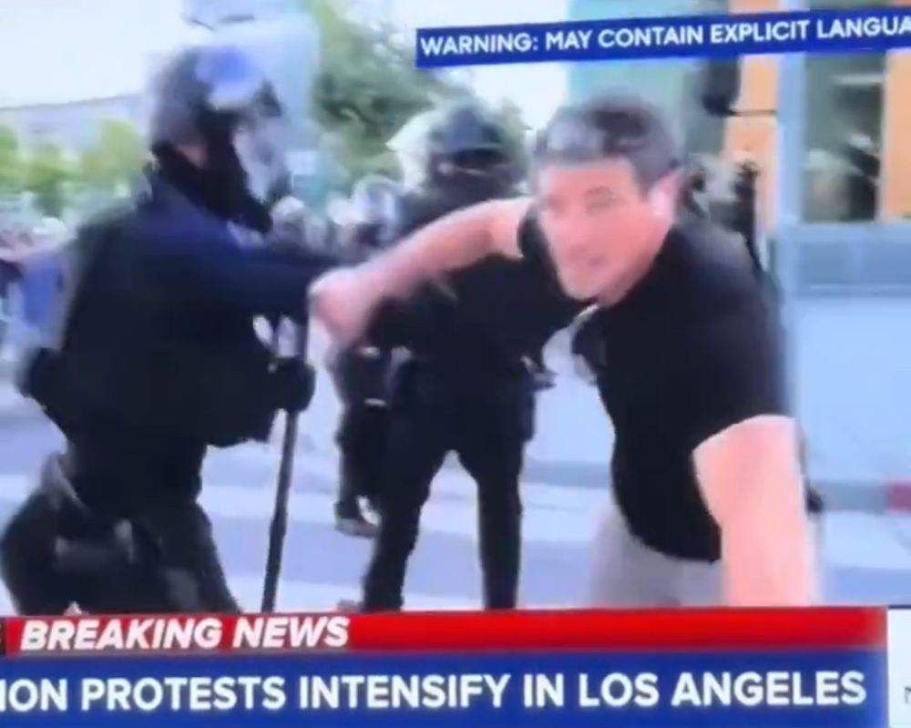 New correspondent holding a LA police officer’s arm in response to being moved when not responding to a move back order.