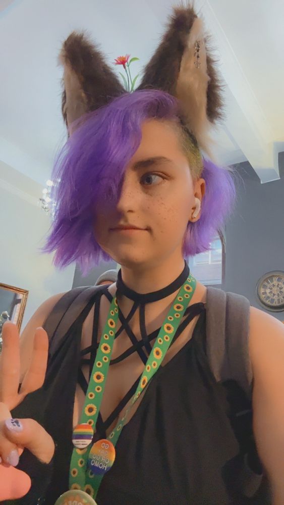 Silly ass wolfie Enfield with her purple hair and strappy crop top, with her silly ears atop her head and disabled sunflower lanyard with some gay badges on it, looking awkwardly off to the side in a hotel lobby.