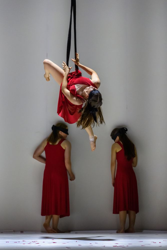 Female aerialist, with long, brown, hair, dressed in red, on a silk, with two blindfolded women in red dresses, behind.
