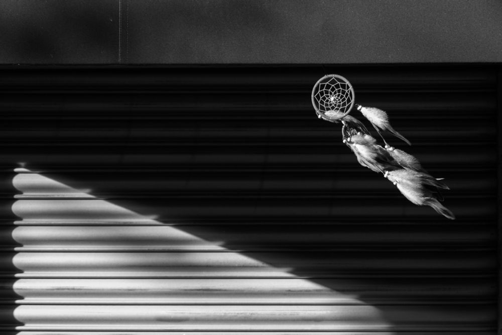 Black and white photo of a dreamcatcher (top right), blowing in the wind, to the right of the frame, against a dark background, with a lighter triangle of light to the bottom left. Honestly, I'd do it better justice if we were both live and interacting!