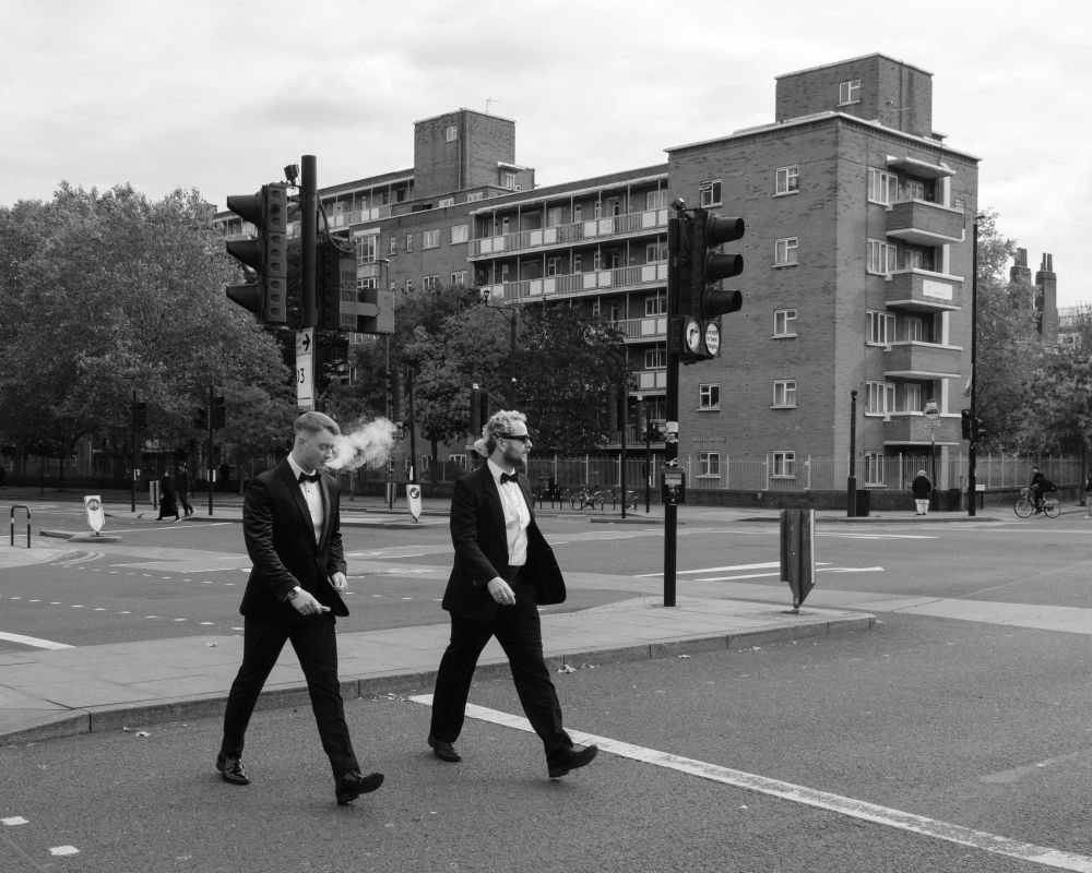 Two young men, in dinner jackets, cross the road, Whitechapel Road, London. There is a block of council flats in the background. The guy on the left is exhaling vape smoke. 