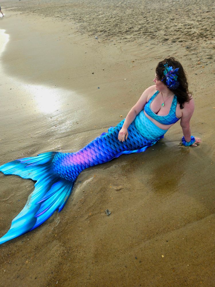 Zippy, a teal, purple, and blue mermaid, sits on a beach looking over to the horizon.
