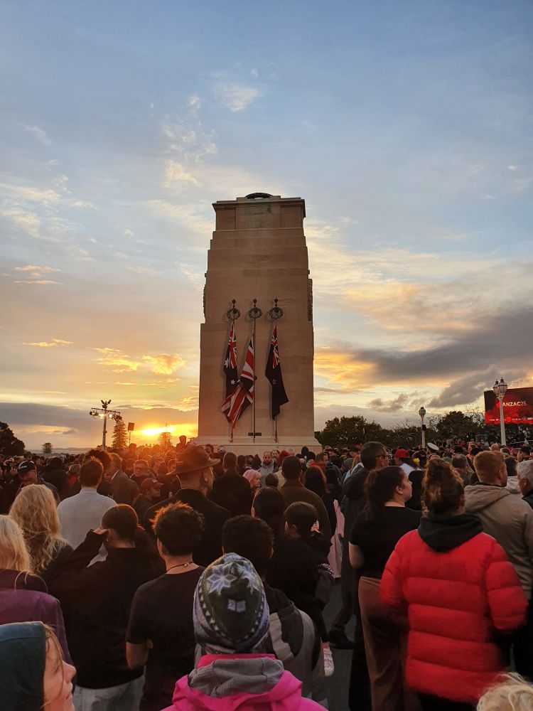 Photograph of a crowd in front of the stone cenotaph at Auckland War Memorial Museum – Tāmaki Paenga Hira. The flags of Australia, New Zealand and the UK hang from poles on the side. The sun is just rising in the background.